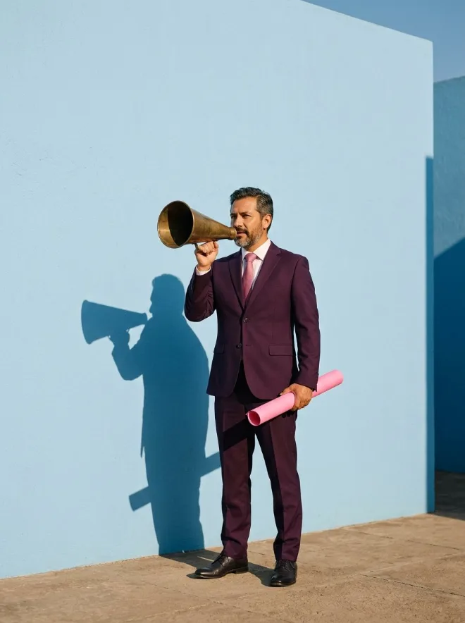 Hombre de traje morado anunciando con un megáfono de latón frente a un muro azul